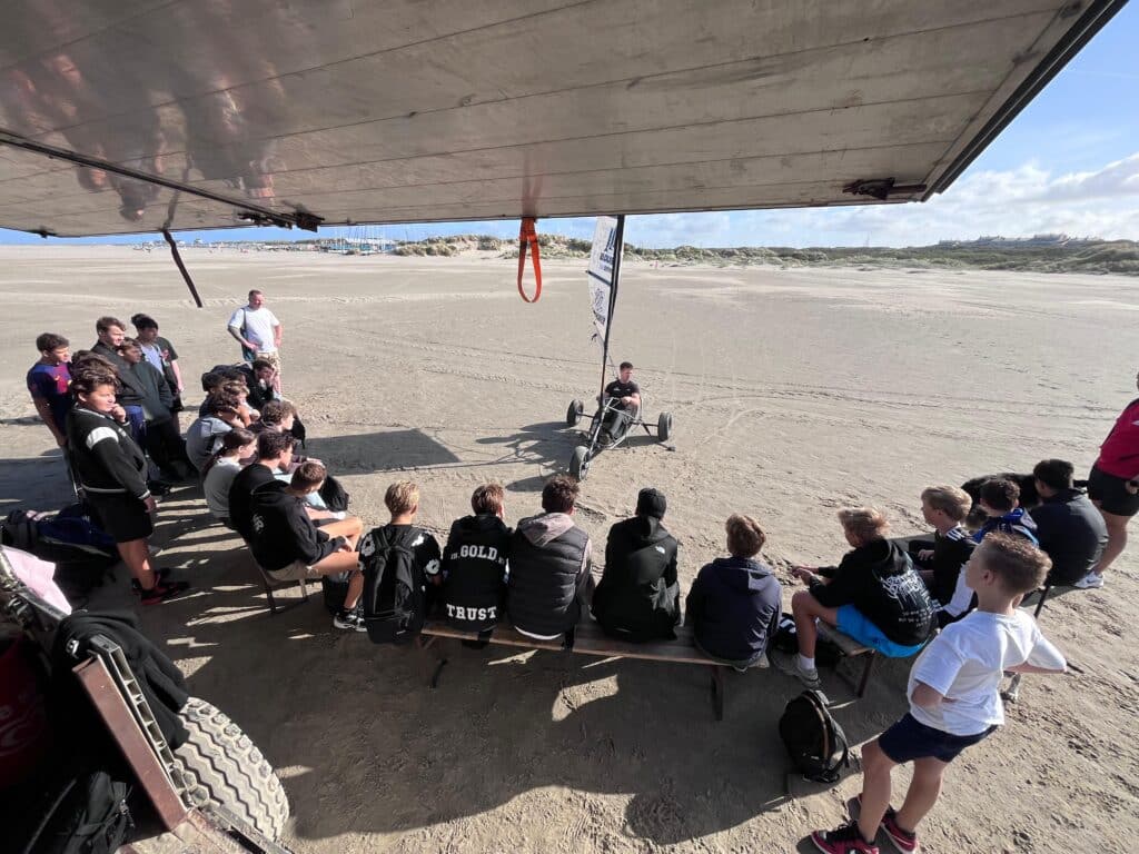 Groep deelnemers luistert naar instructeur bij blokarten IJmuiden met 5,5m zeil op zonnig strand